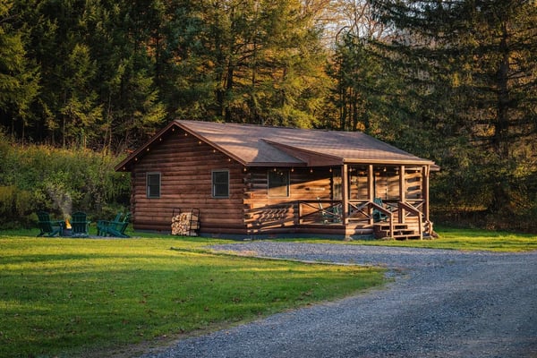 Log cabin with covered porch and Adirondack chairs, surrounded by autumn forest and gravel driveway