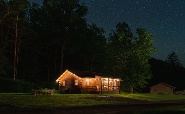 Illuminated log cabin in forest clearing at night with starry sky and tall trees surrounding property